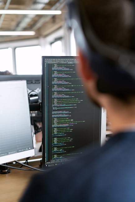 The image shows a person with short dark hair viewed from behind, working at a desk in an office environment. They are focused on a computer screen displaying lines of colorful code, indicating progra