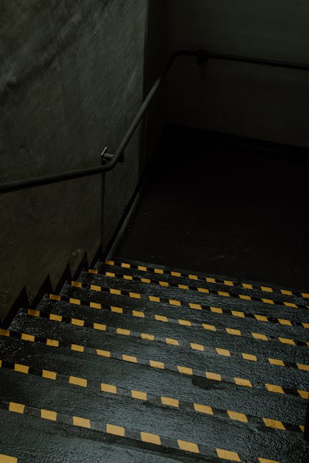 A downward view of a residential stairwell with black-painted steps that have yellow and black striped anti-slip tape along the edges. The walls are dark, textured concrete, and a metal handrail runs 
